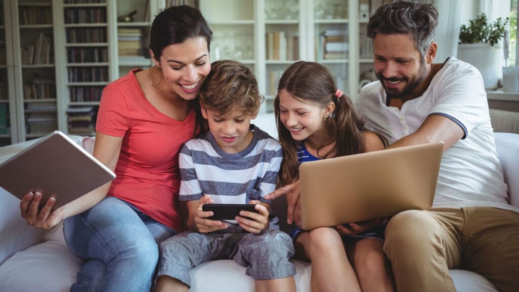 Photo of a family sitting on a couch using a laptop, phone, and tablet