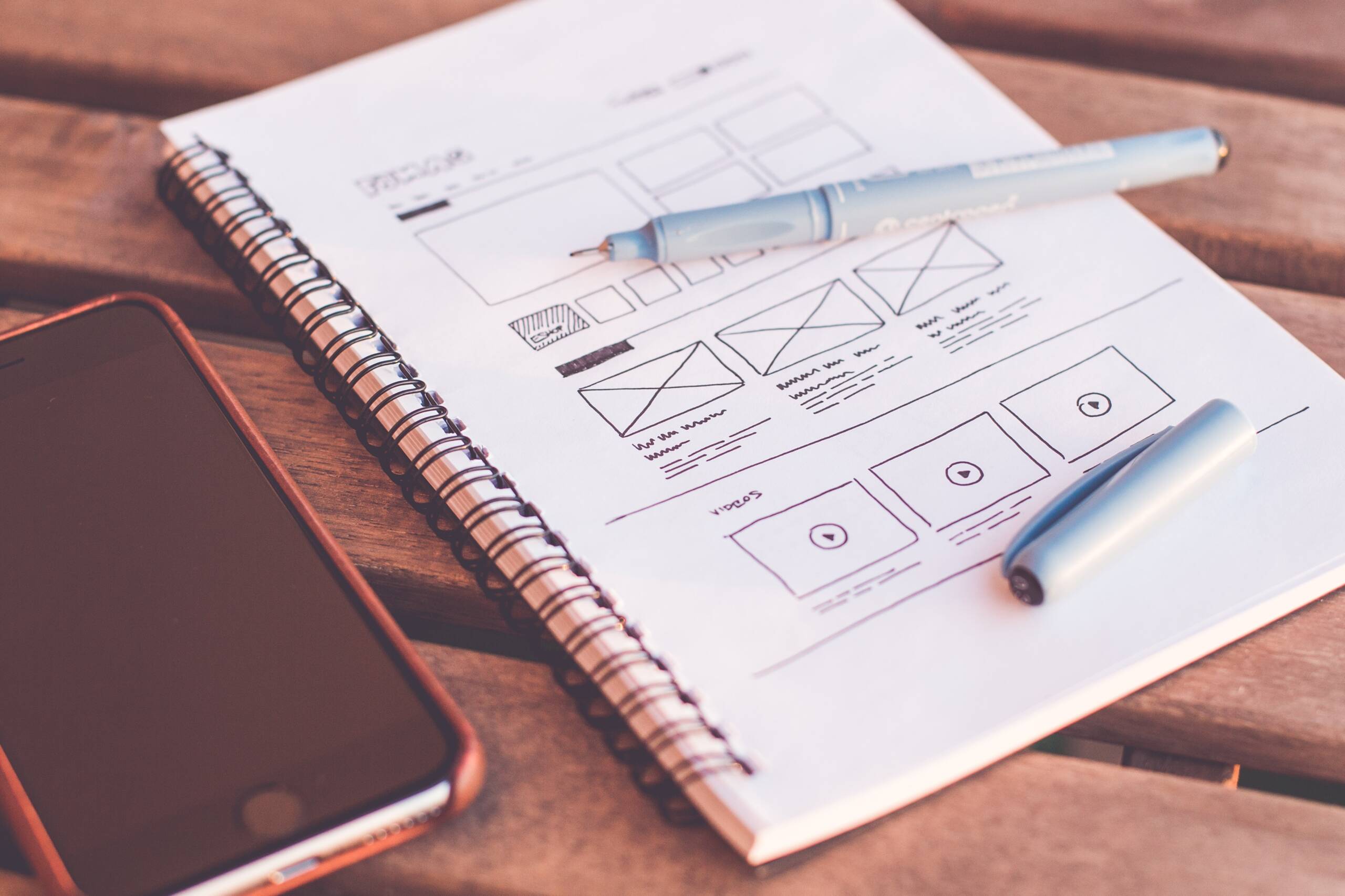 Notebook with UX wireframe sketches, a pen, and a smartphone on a wooden table, representing the planning phase of a UX design assessment.