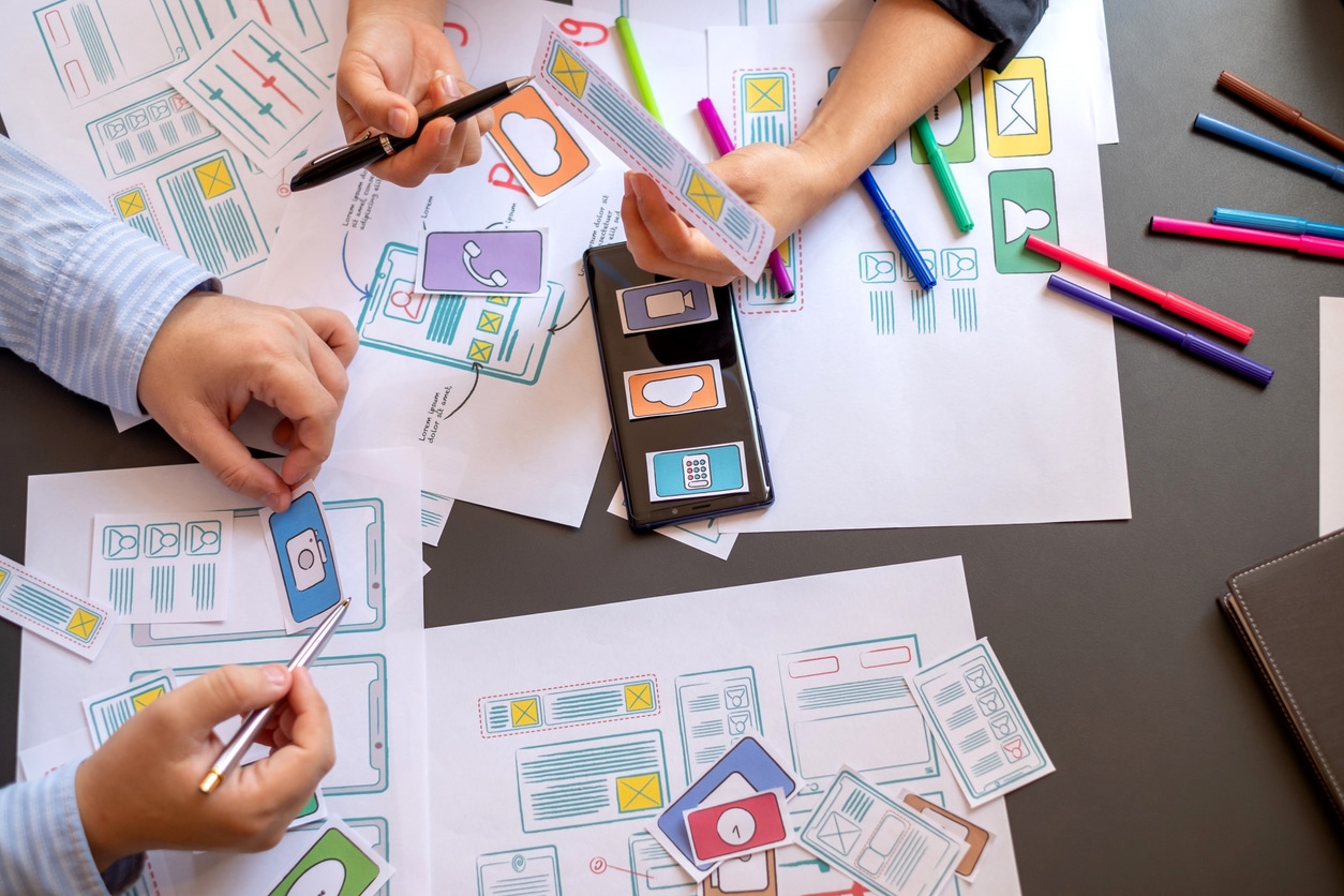 Hands of designers working on mobile UX wireframes, using paper cutouts and sketches to prototype an application interface.
