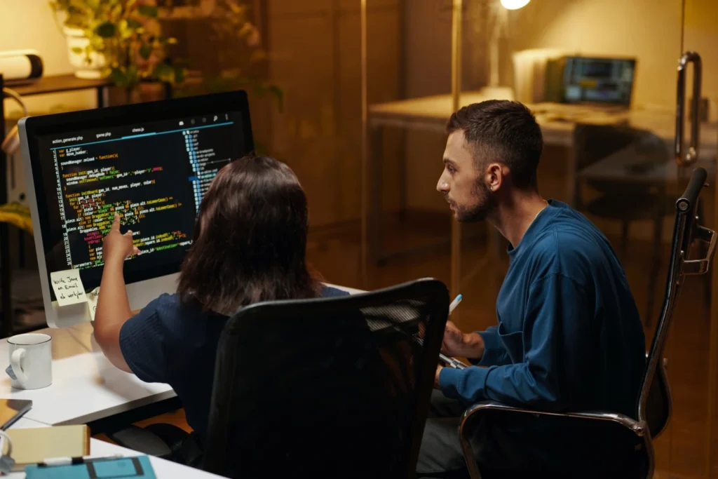 Two software engineers reviewing source code on a large monitor and pointing out bugs during a QA session.