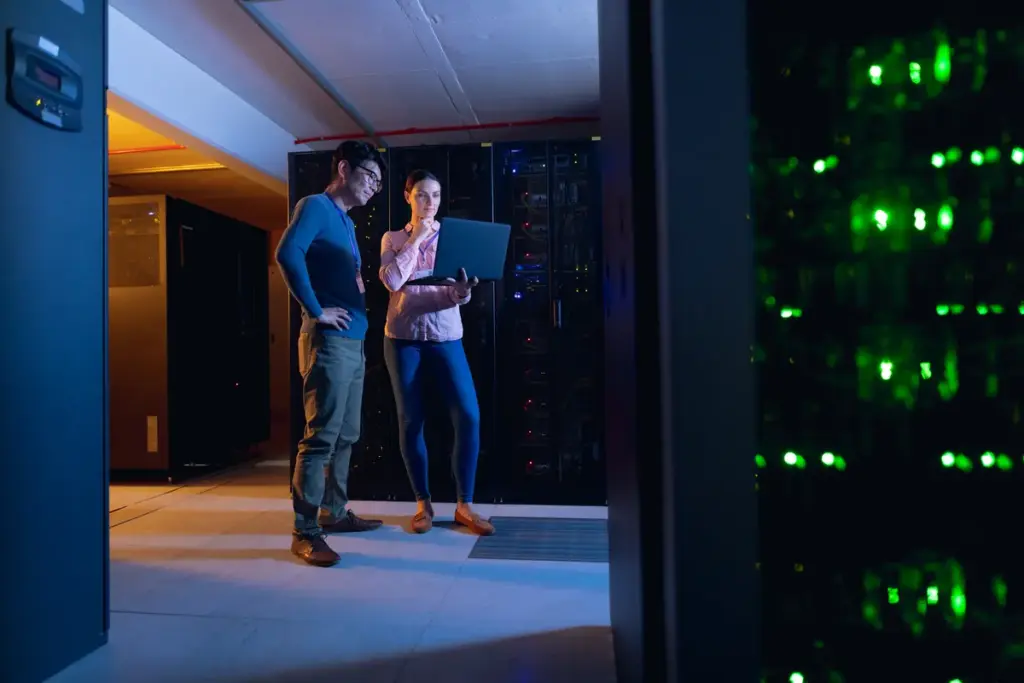 Two cloud infrastructure engineers reviewing disaster recovery systems on laptop in modern data center with server racks showing active network connections
