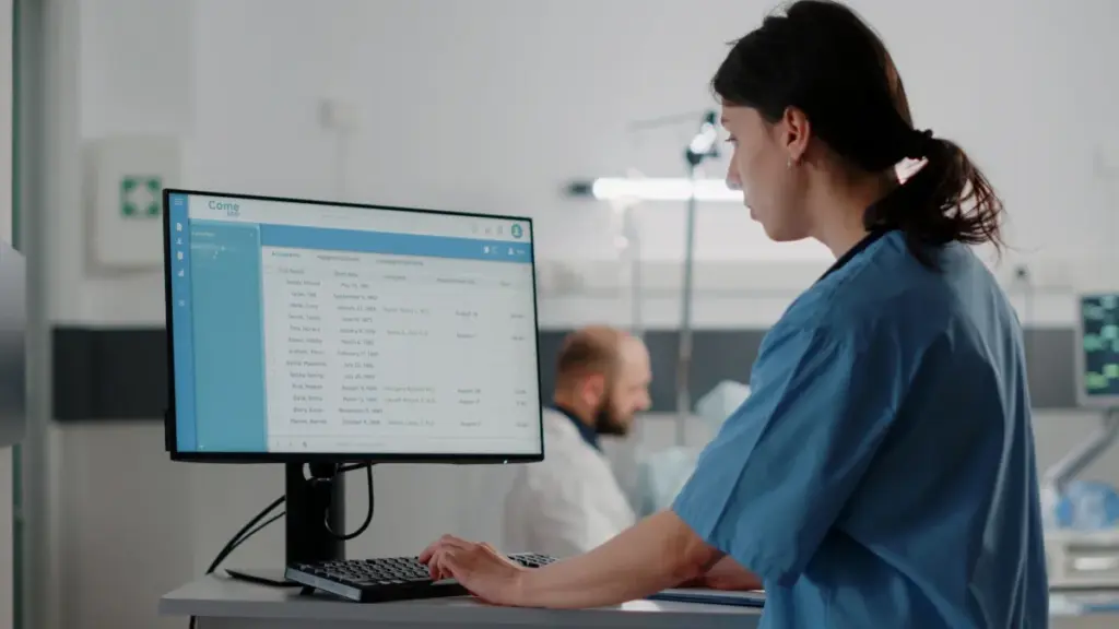 A healthcare professional in blue scrubs using a patient management software system on a computer, representing unosquare's engineering solutions for the healthcare industry.